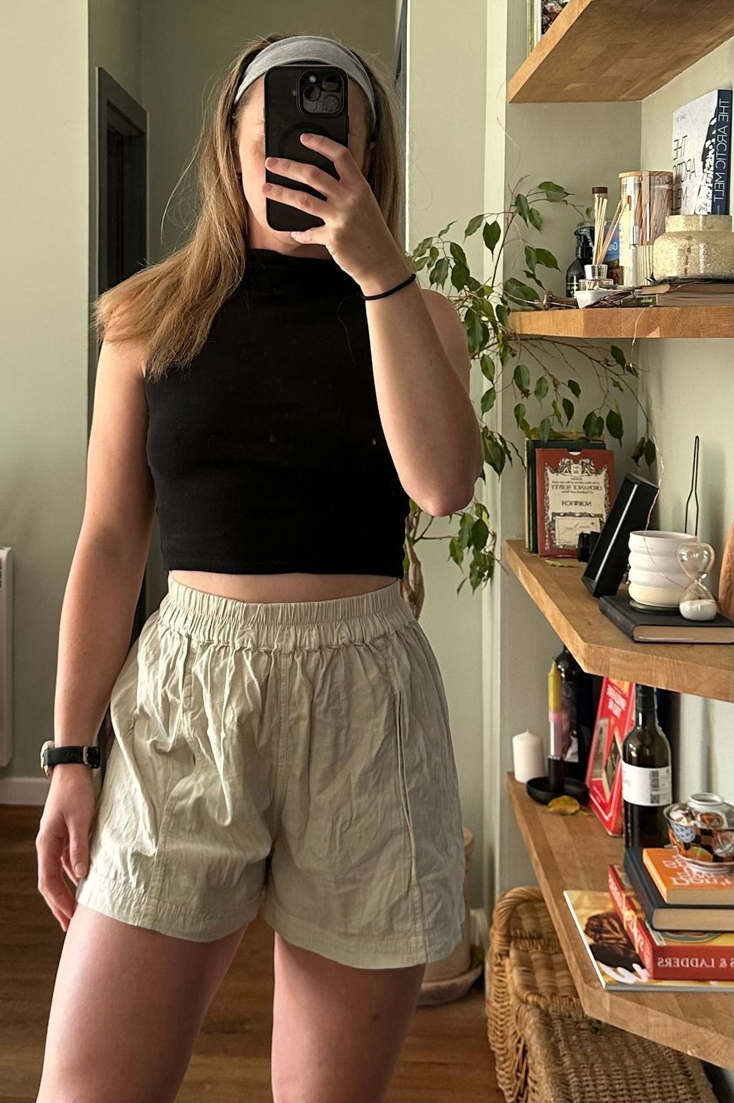 A woman is taking a selfie in a room with shelves and various items, including a plant and books.